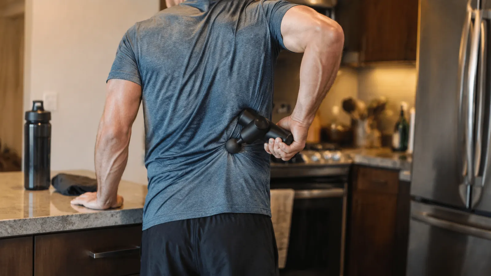 Person using a percussion massage gun on their lower back while standing at a kitchen counter, suggesting a quick recovery routine at home