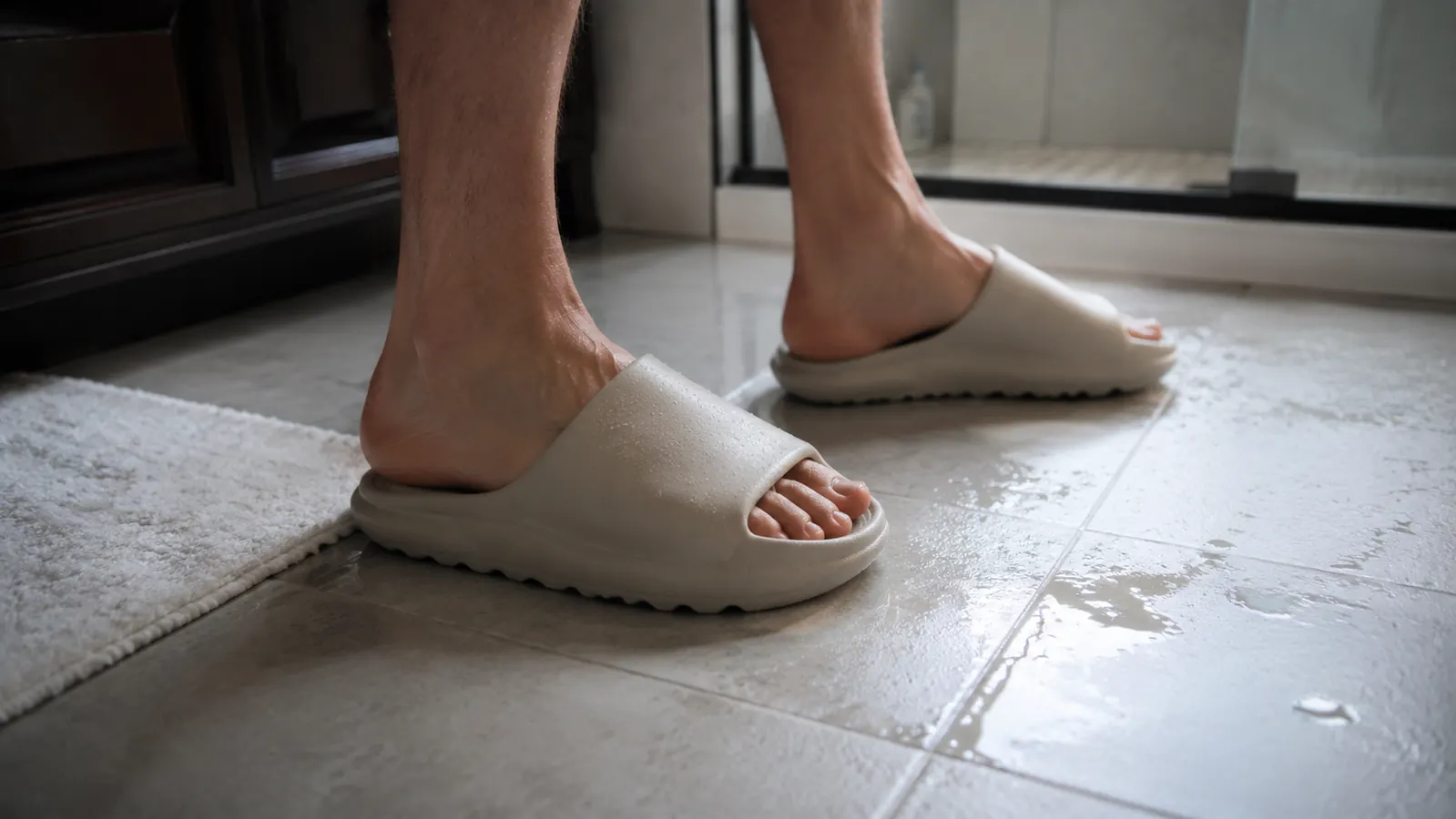 Close-up of feet wearing thick recovery slides stepping cautiously toward wet bathroom tile, illustrating the traction caution on slick floors