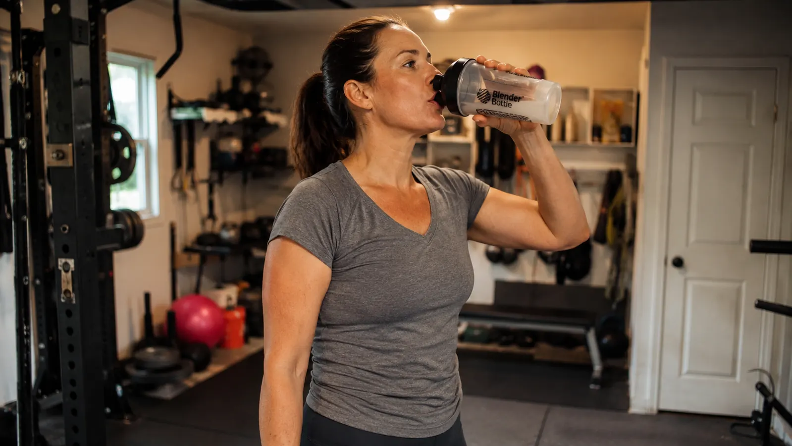 A woman in her late thirties drinking from a shaker bottle in a home gym, wearing casual workout clothes, expression calm and matter-of-fact
