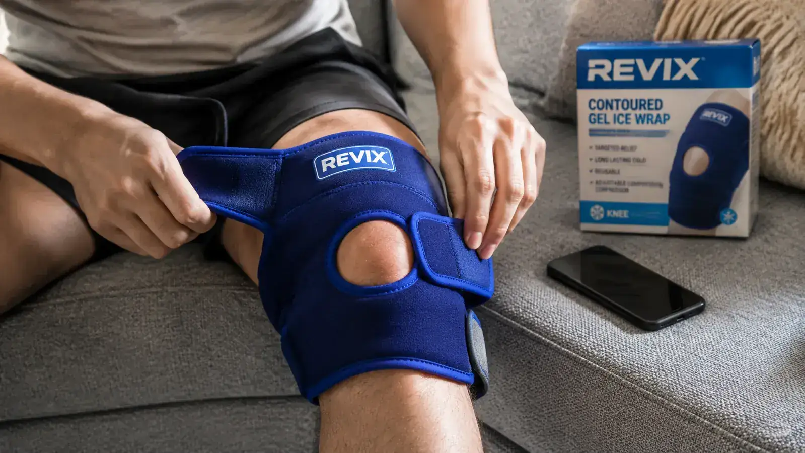 Person wrapping a blue gel ice pack around their knee while seated on a couch at home, phone on the cushion beside them